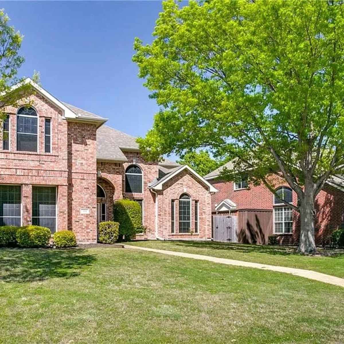 A two-story brick house with a landscaped front yard and large windows is situated on a green lawn.