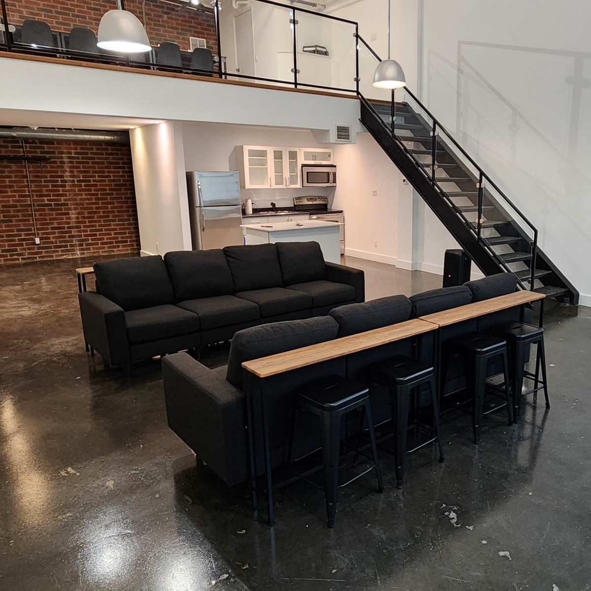 A spacious, modern loft features a black sectional sofa with a wooden tabletop, a metal staircase, and a kitchen area visible in the background.