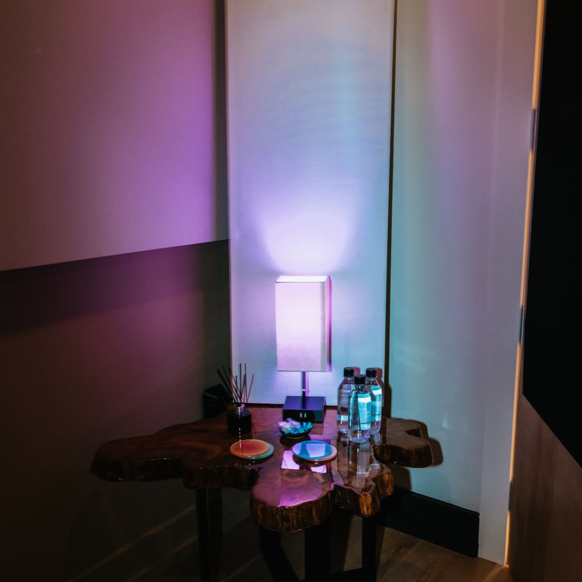 A small corner table made of natural wood with a lamp, bottled water, and decorative items against a softly lit wall.