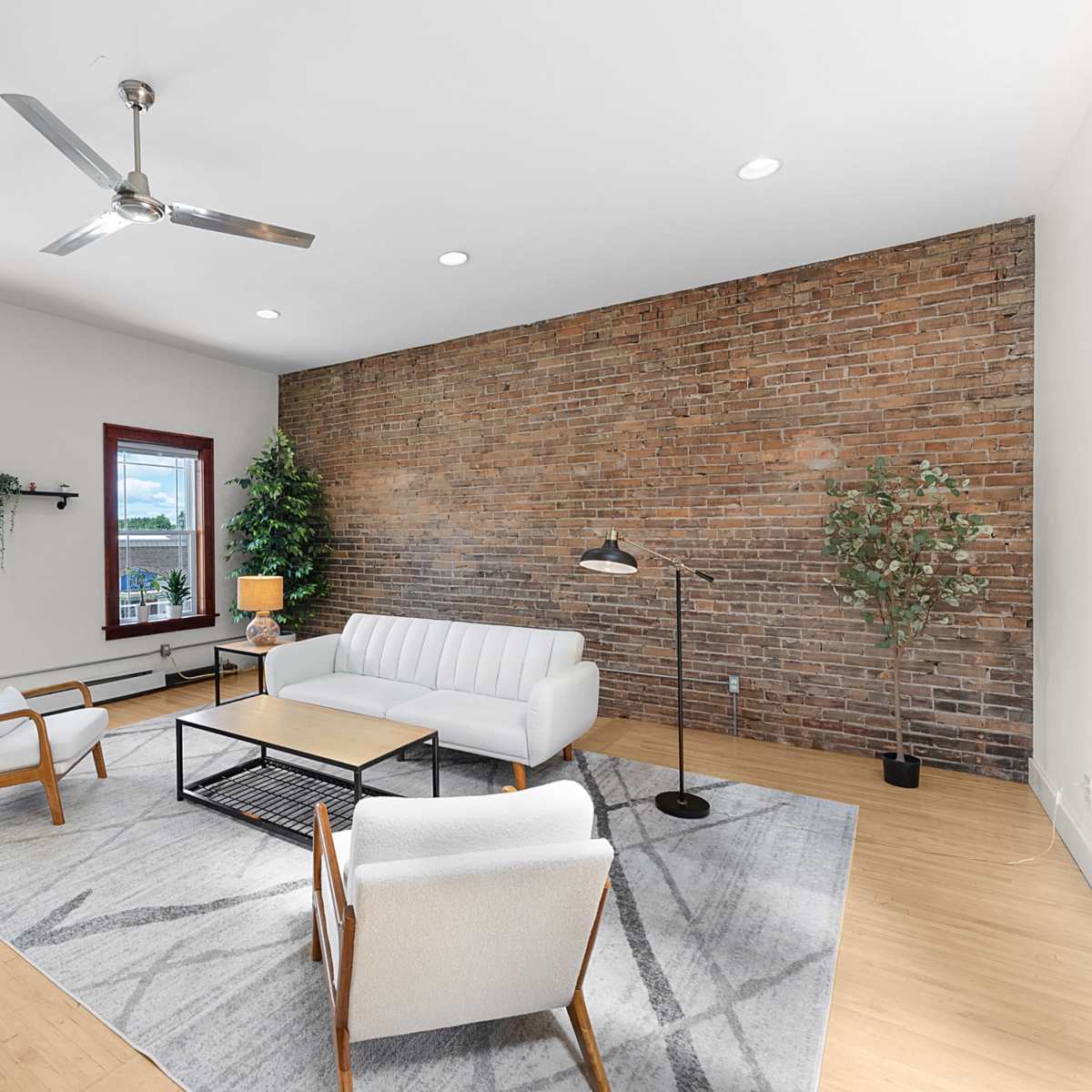 The room features a modern living space with a white sofa, wooden chairs, a coffee table, and exposed brick walls.