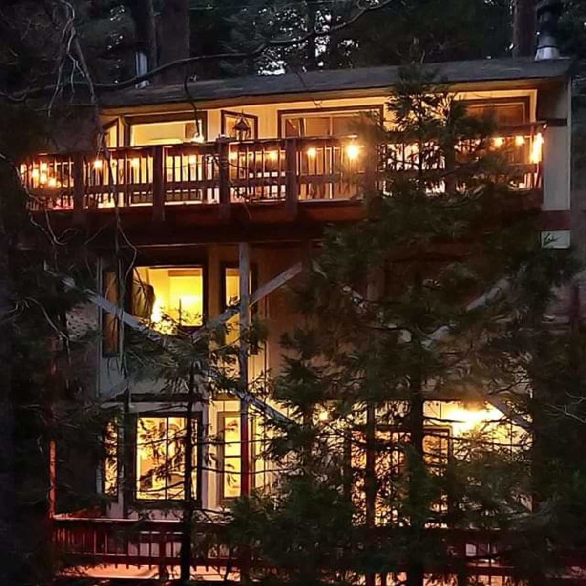 A two-story cabin with large windows and multiple balconies is nestled among tall trees in a forested area at dusk.