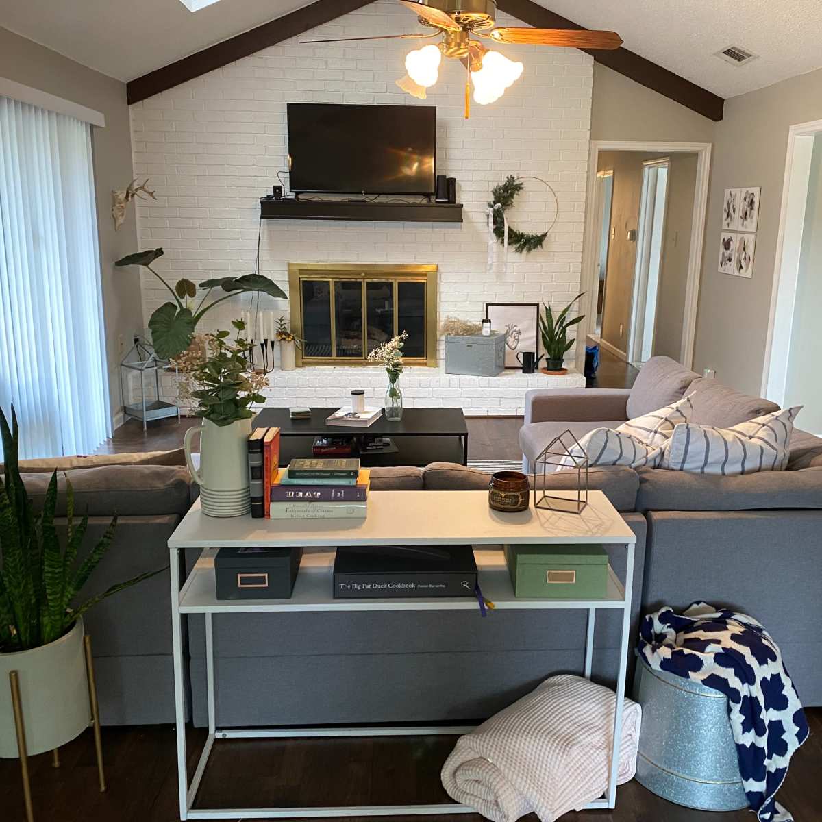 The living room features a gray sectional sofa facing a fireplace, with a coffee table in front and plants adorning the space.