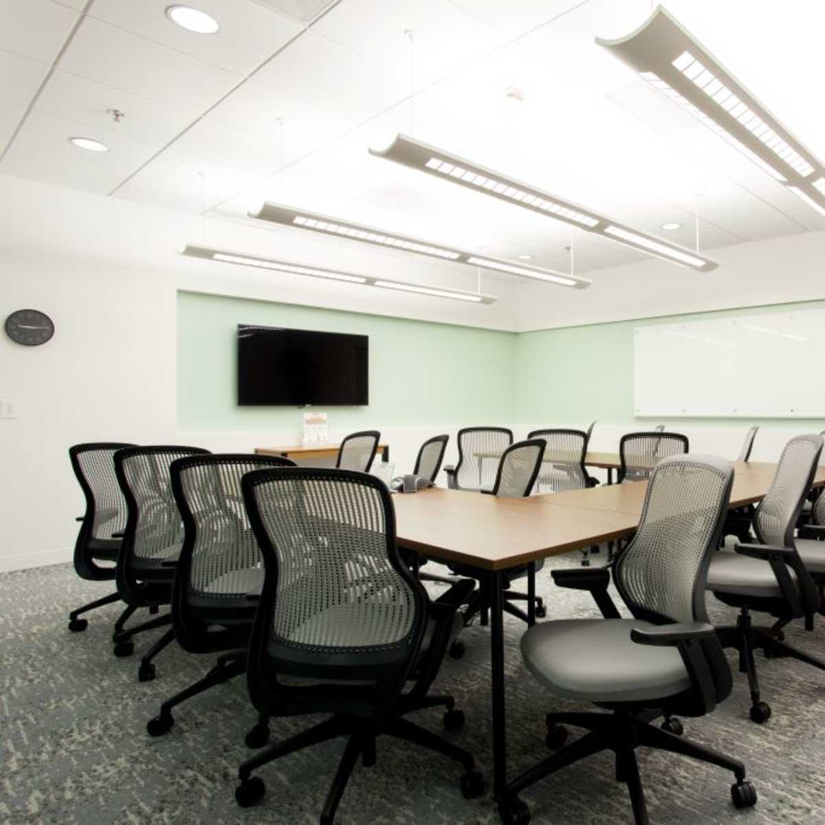 A conference room features a large wooden table surrounded by ergonomic chairs, with a TV and whiteboard on the walls.