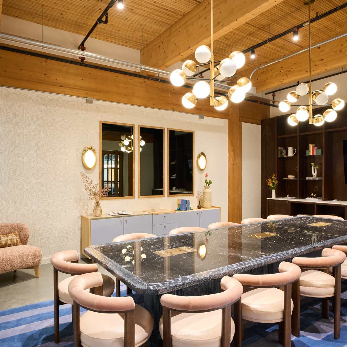 A modern dining area with a large black marble table surrounded by light-colored chairs, complemented by a patterned rug and contemporary lighting fixtures.