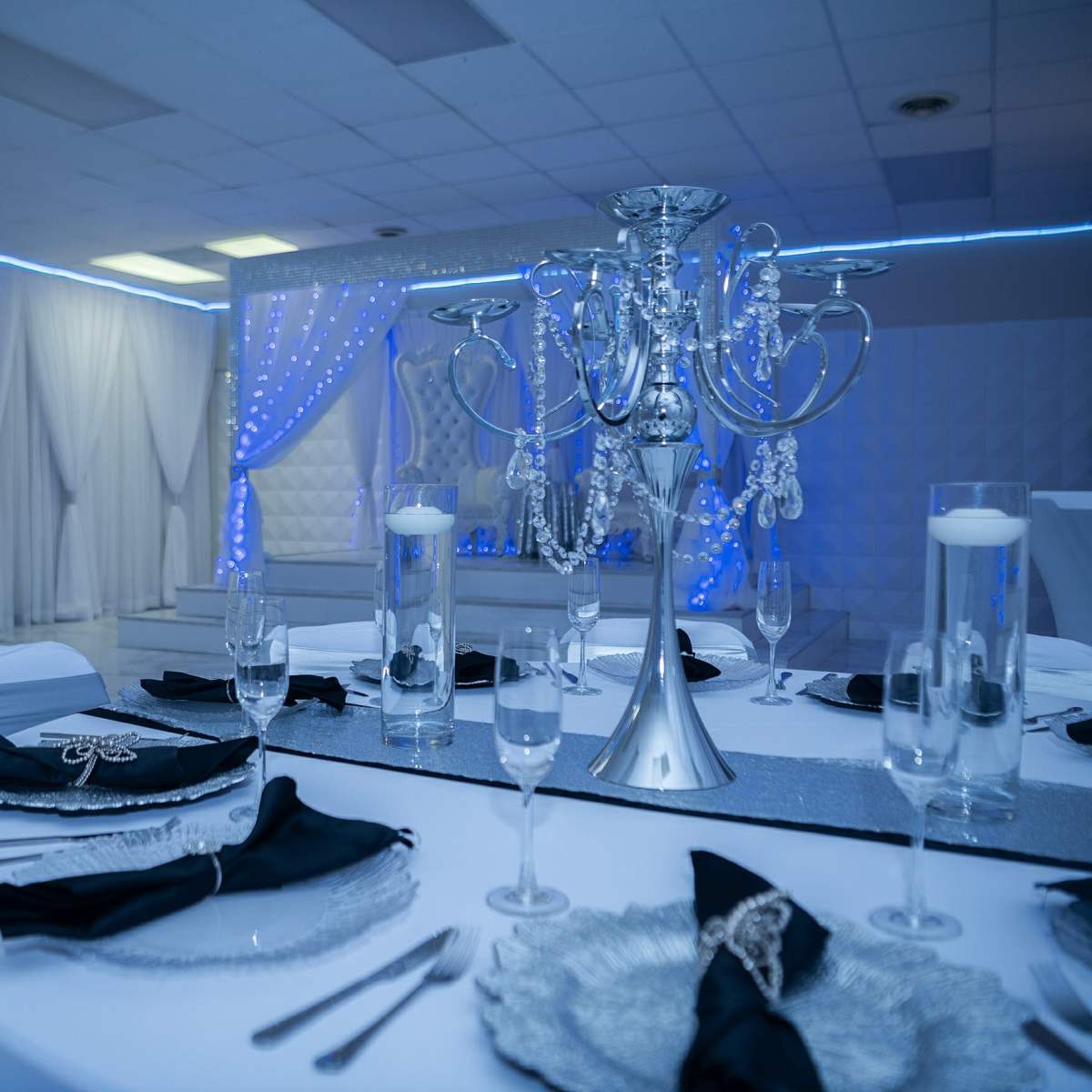 A elegantly decorated dining area with a round table set with black napkins, silver tableware, and a large silver candelabrum as the centerpiece, surrounded by white chairs and blue lighting.