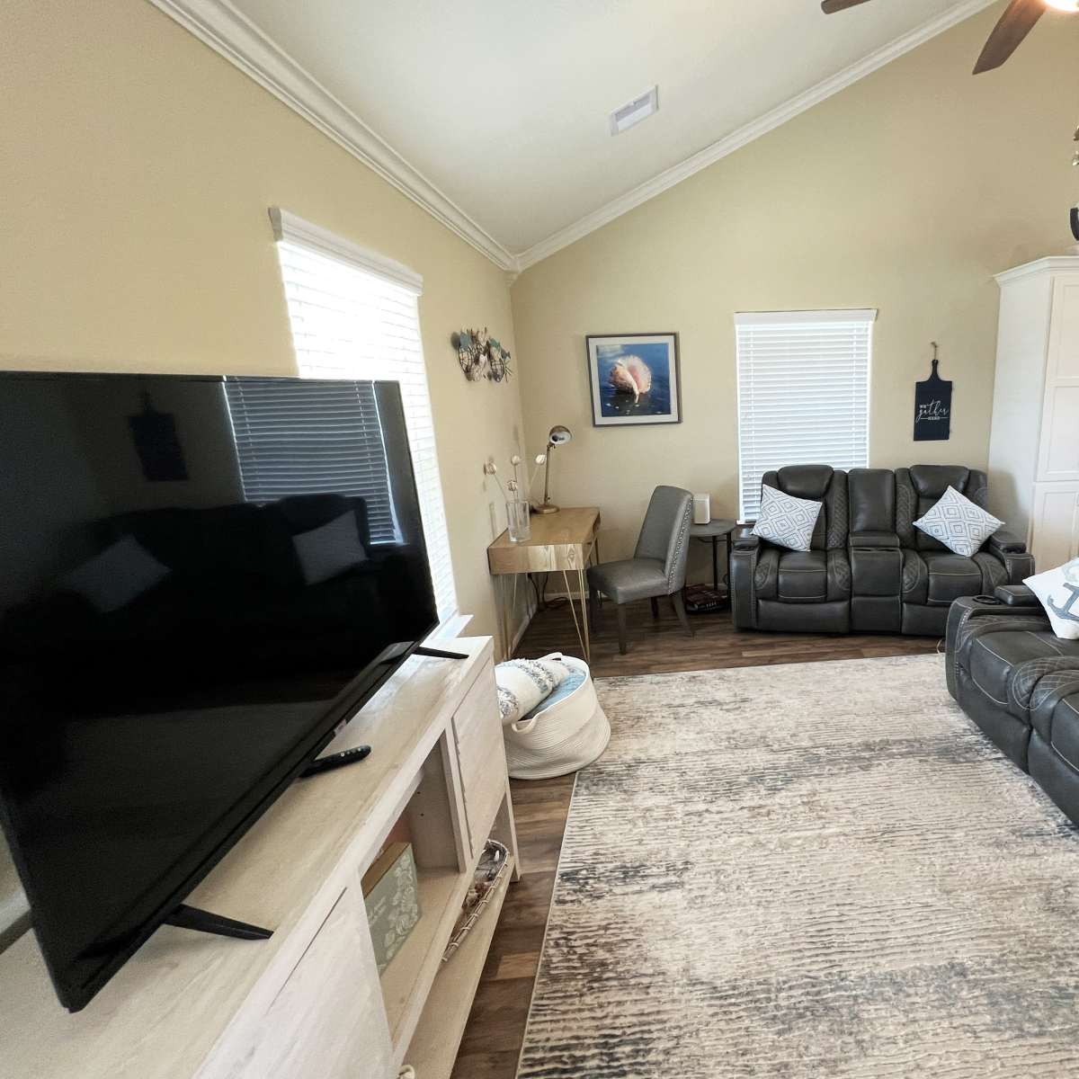 The living room features a large flat-screen television, two reclining sofas with decorative pillows, and a desk positioned near windows, set on a patterned area rug.