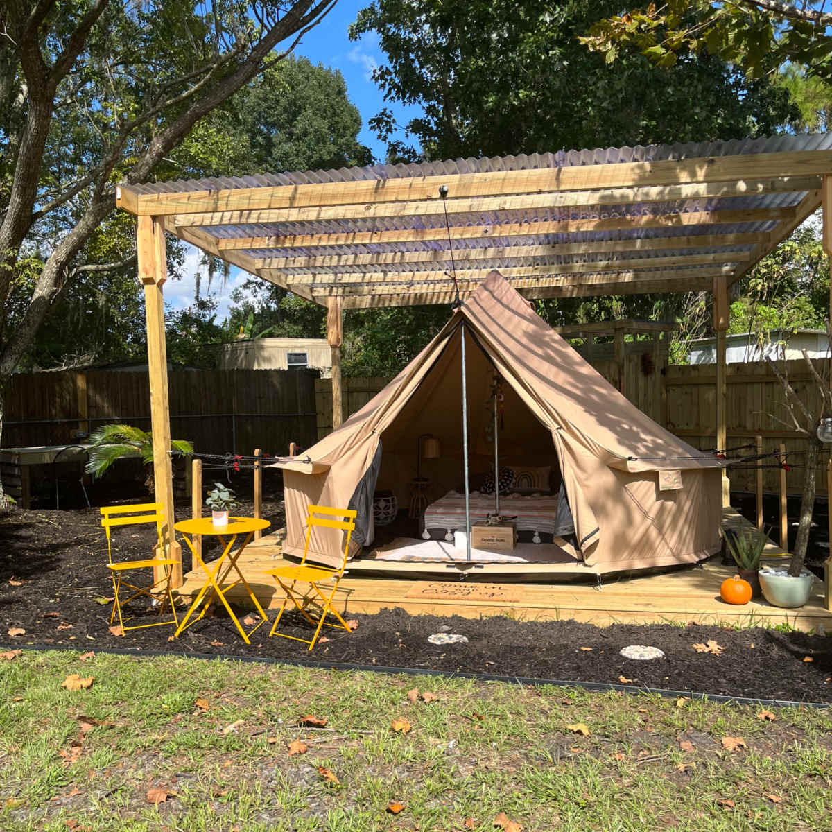 A tent is set up under a wooden pergola, accompanied by yellow chairs on a grassy area, with trees and a fenced background.