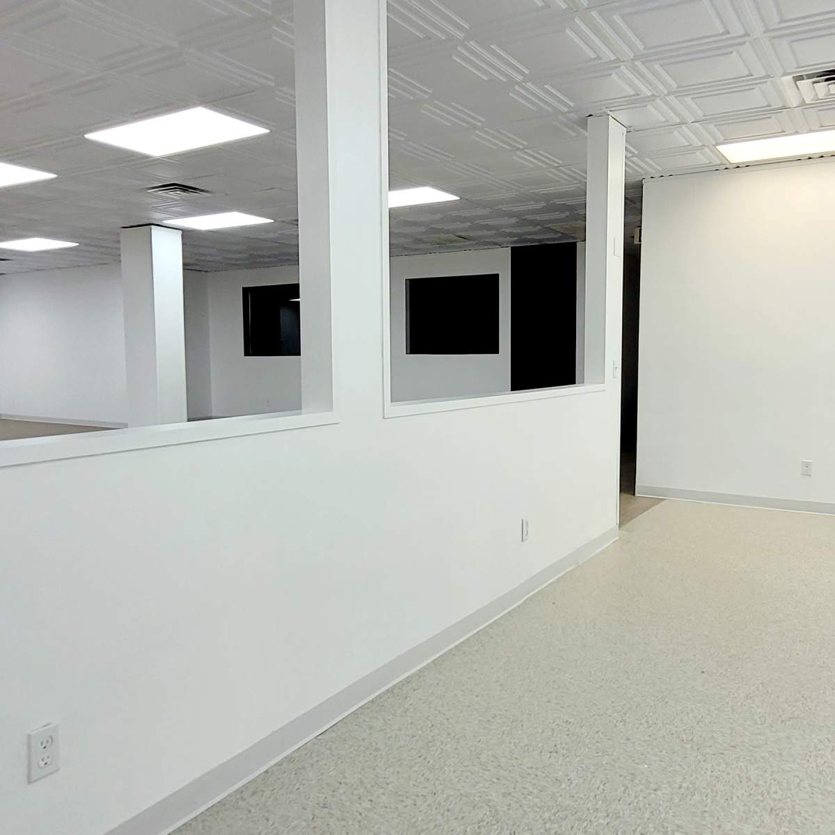 An empty room with white walls, a ceiling with tile patterns, and a light-colored floor, featuring large fluorescent lights and a window that opens to another space.