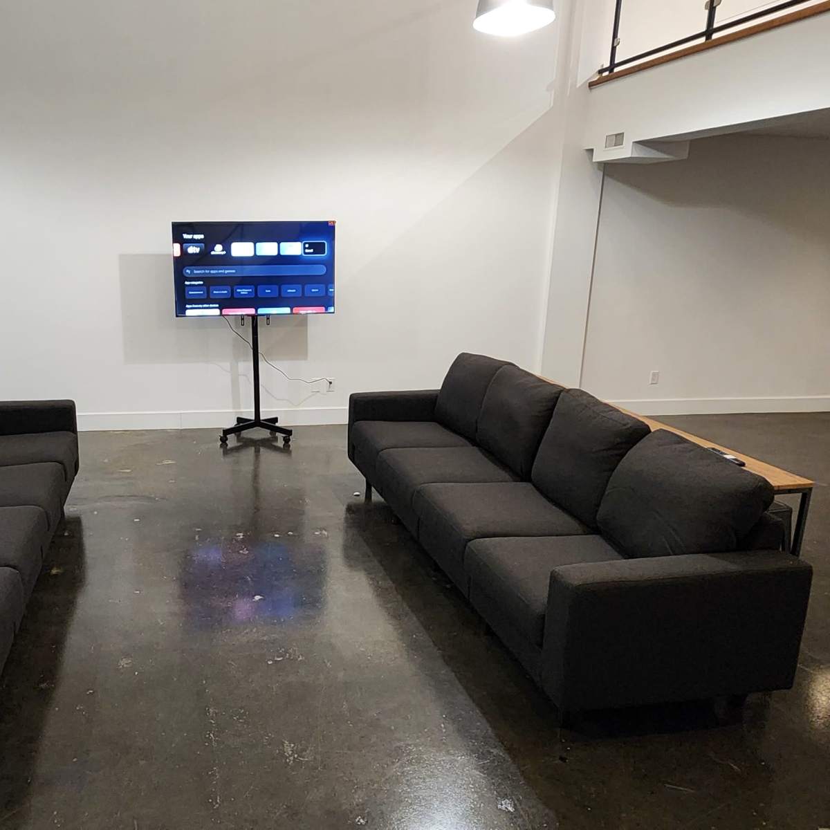 Two black sofas facing a mounted television in a spacious room with a polished concrete floor and white walls.