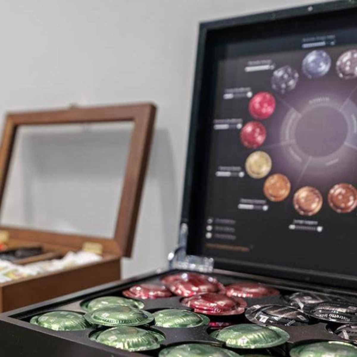 A display of coffee and tea pods in a wooden box alongside utensils and a set of white cups in the background.