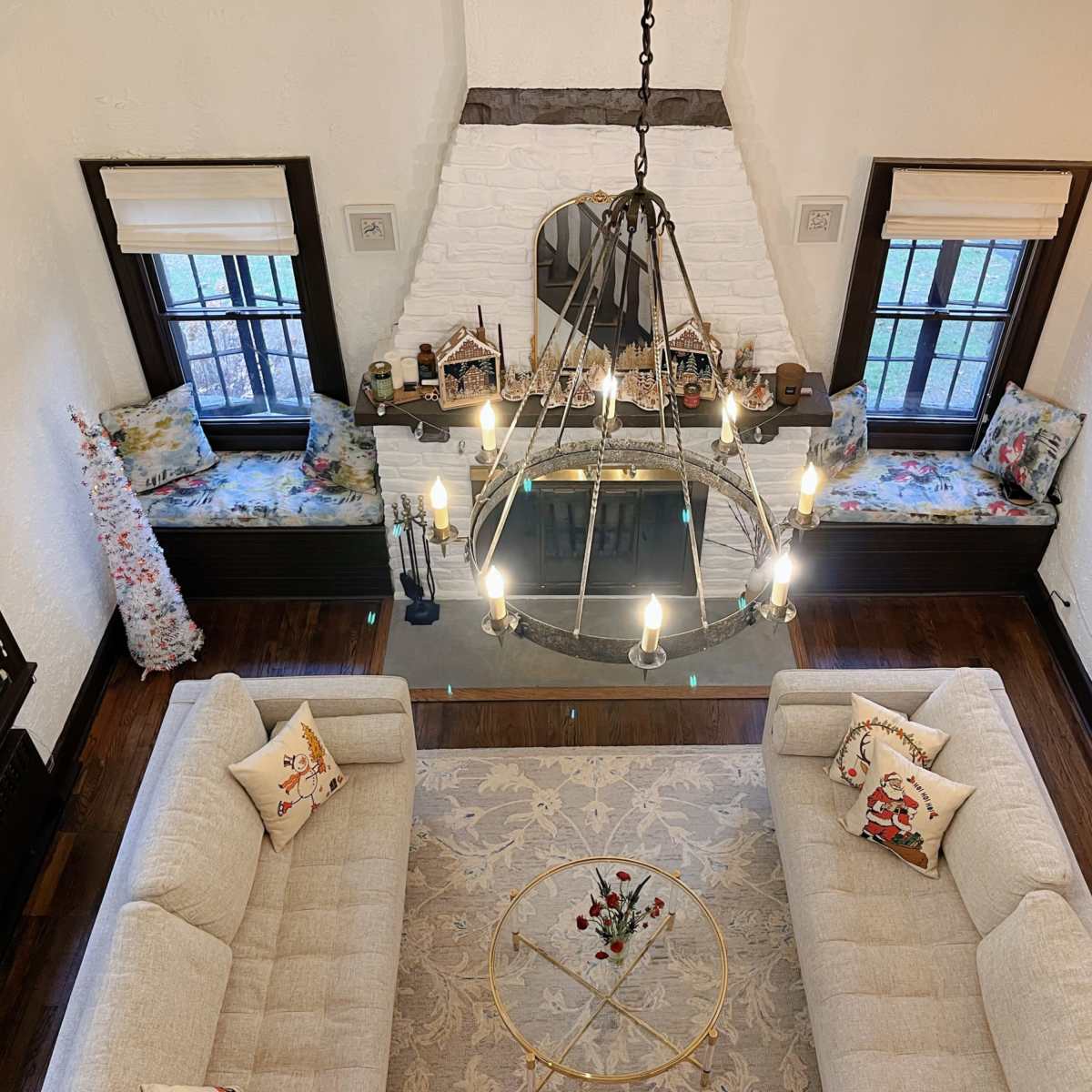 A living room with a high ceiling, featuring a chandelier, two light-colored sofas, a round coffee table, and a white brick fireplace.