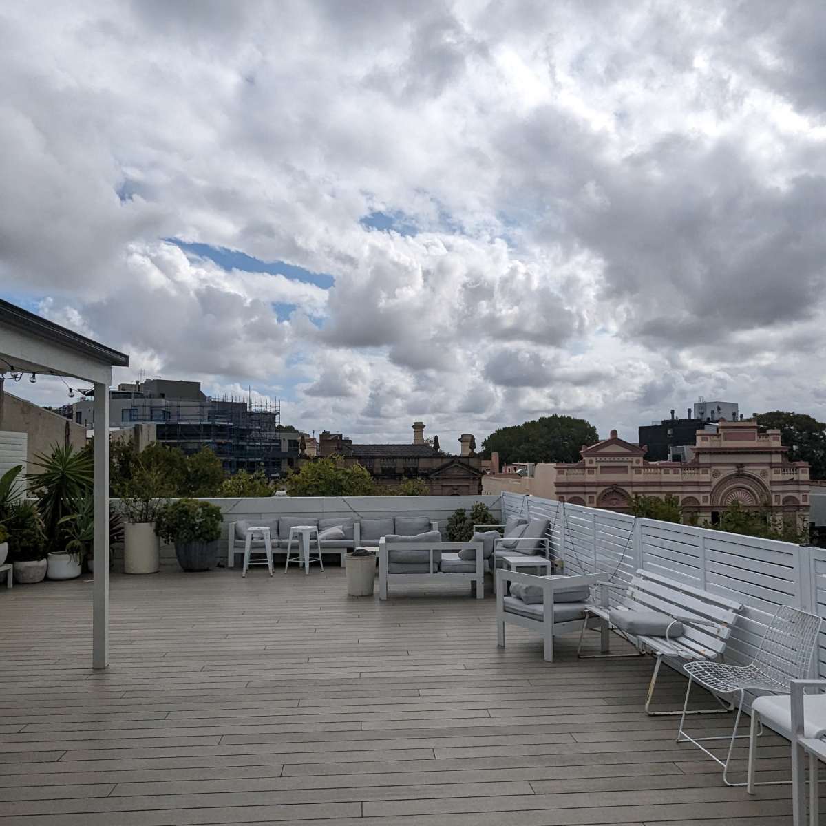 A rooftop terrace with white chairs and tables, surrounded by cloudy skies and a view of buildings in the background.
