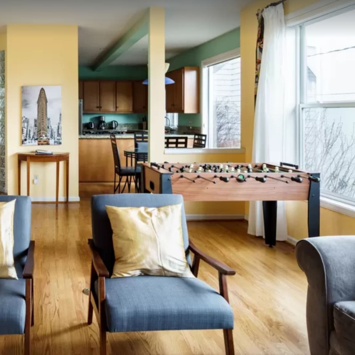A living room with two chairs facing a wooden table, a foosball table, and a kitchen area in the background.