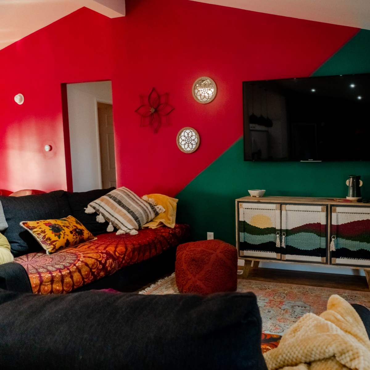 A living room features a dark sofa, colorful accent walls, a television mounted above a wooden cabinet, and decorative items like a rug and plants.