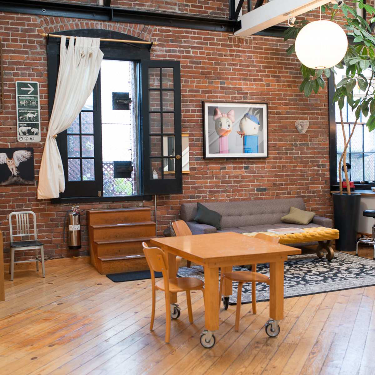 A modern loft-style living space with exposed brick walls, a large window, a gray sofa, a wooden dining table, and a small kitchen area.