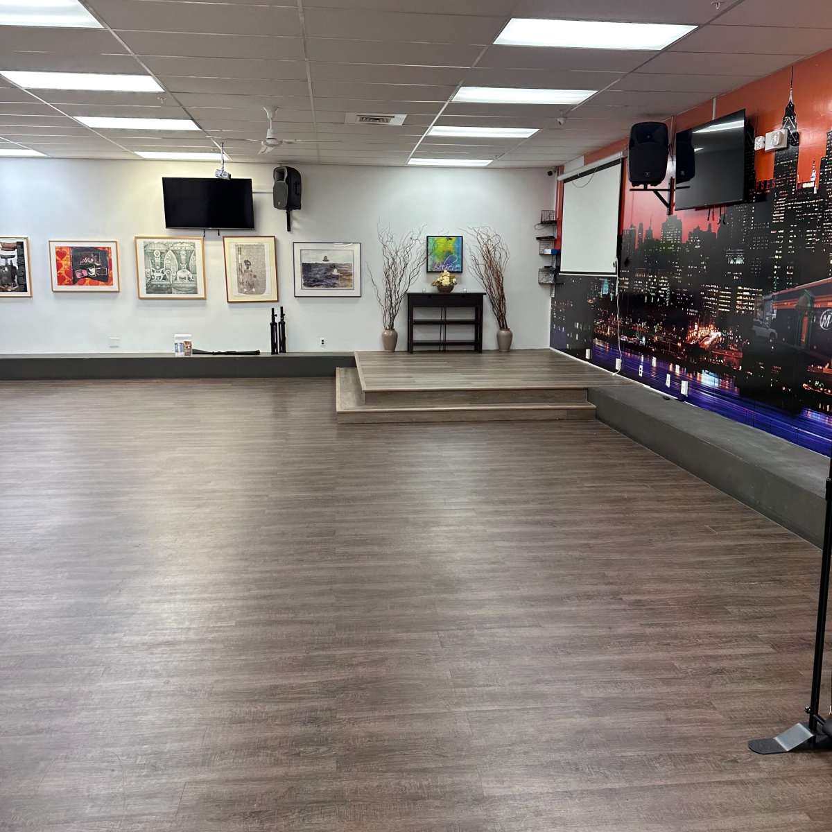 A spacious indoor room with a wooden floor, a small elevated stage at one end, wall-mounted televisions, and artwork hanging on the walls.