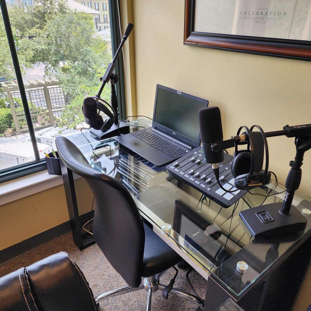 A glass desk in a well-lit room features a laptop, audio equipment, microphones, and headsets, with a view of greenery outside the window.