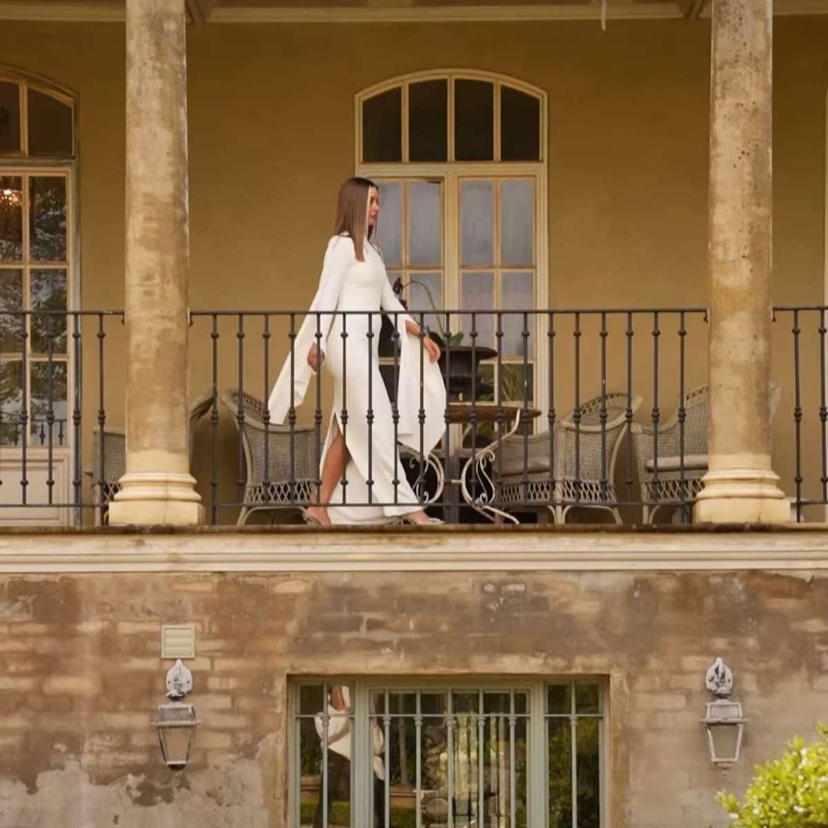 A woman in a white dress walks along the balcony of a two-story building with large windows and decorative plants.