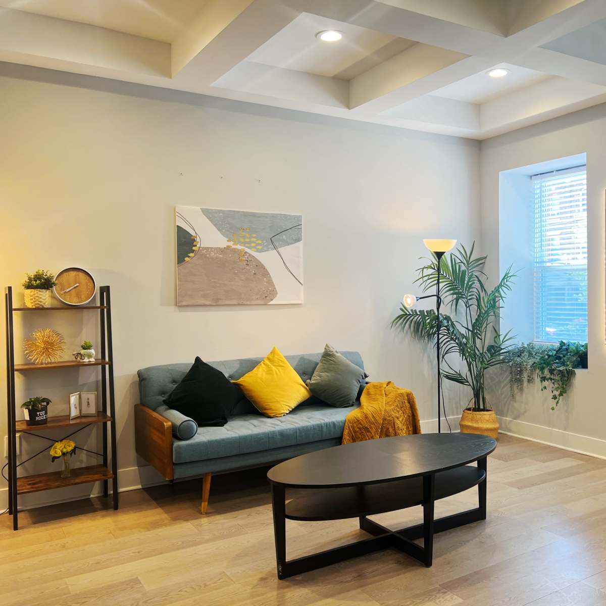 A modern living room features a teal sofa with yellow cushions, a black coffee table, a bookshelf with decorative items, and large windows allowing natural light to illuminate the space.