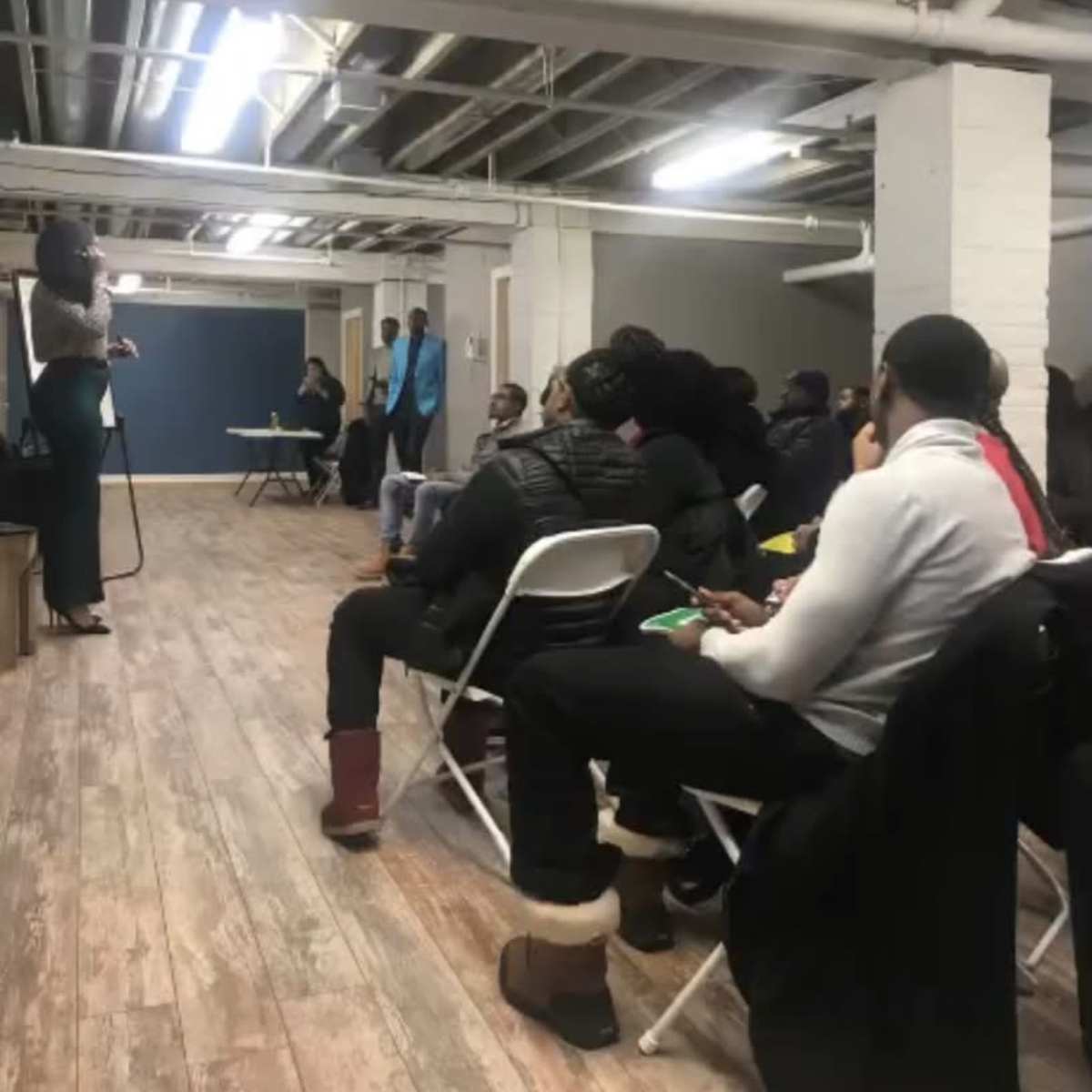 A speaker stands in front of an audience seated in chairs at a gathering in a basement setting.