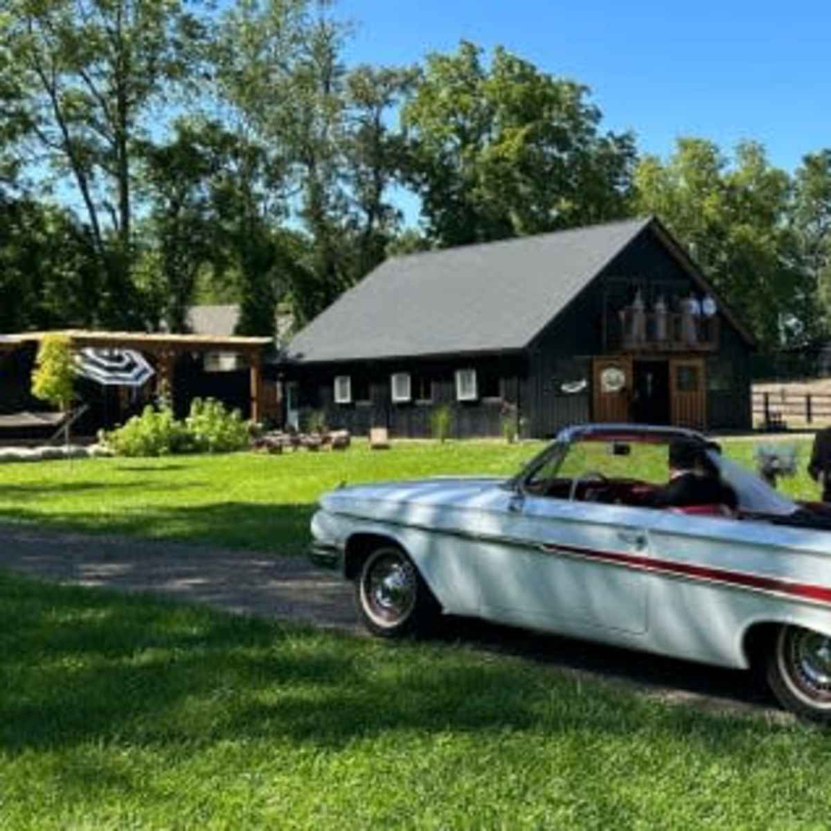 A classic convertible car is parked on a gravel path in front of a black barn while two individuals stand beside it on a lush green lawn.