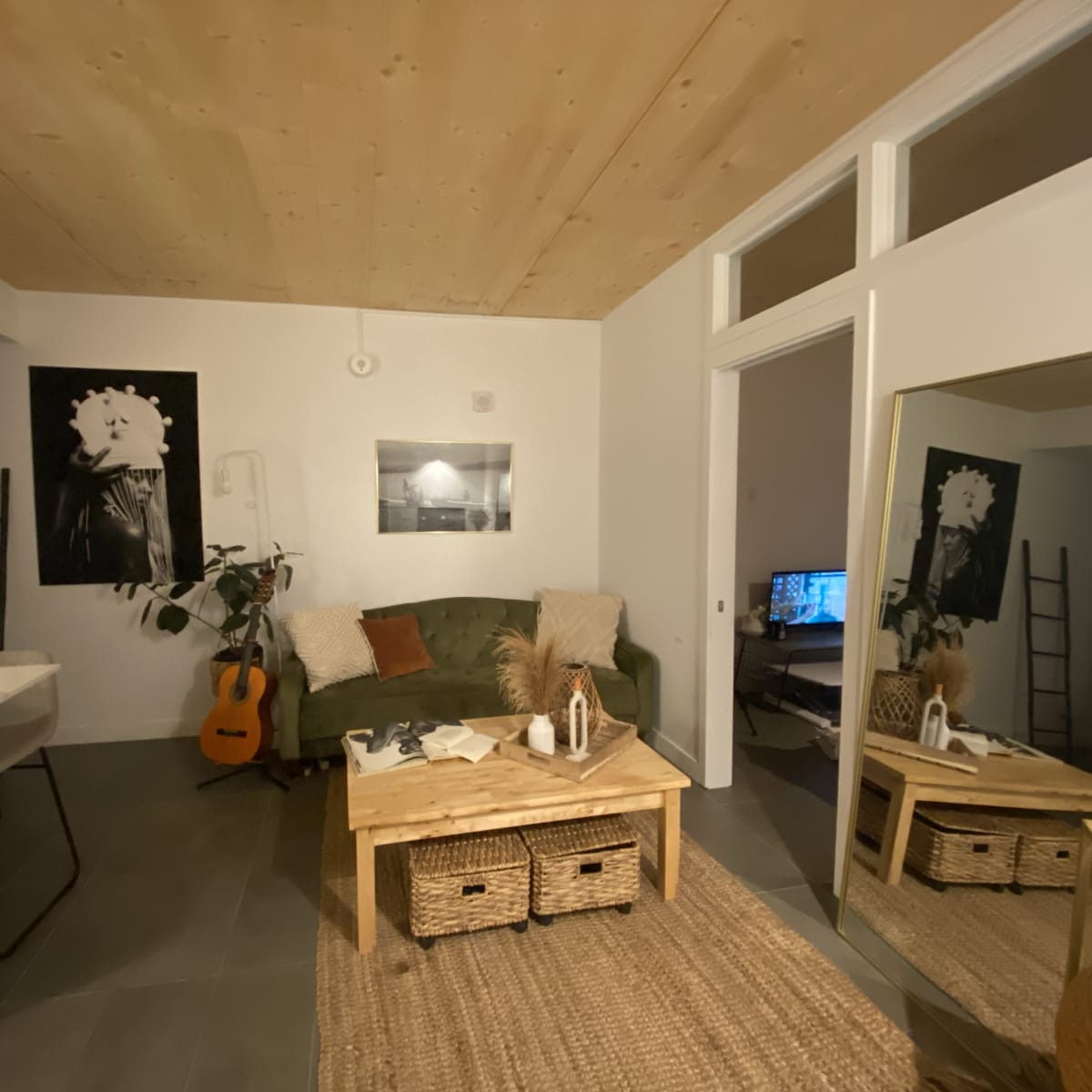 A modern living room with a green couch, a wooden table, and a mirror reflecting a workspace with a computer.