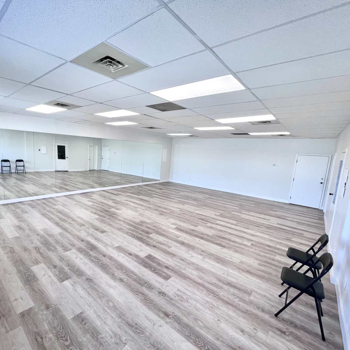 A spacious, empty room with a wooden floor, large mirrors on one wall, and two chairs placed against another wall.