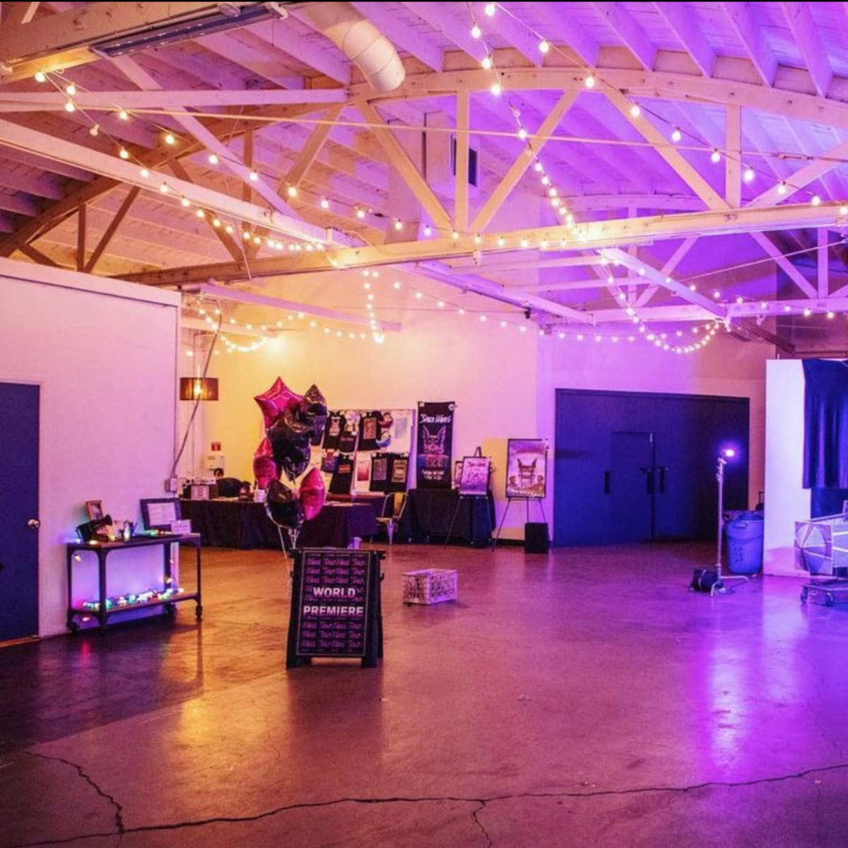 A spacious event venue with string lights overhead, decorated with colorful balloons and banners for a special event.