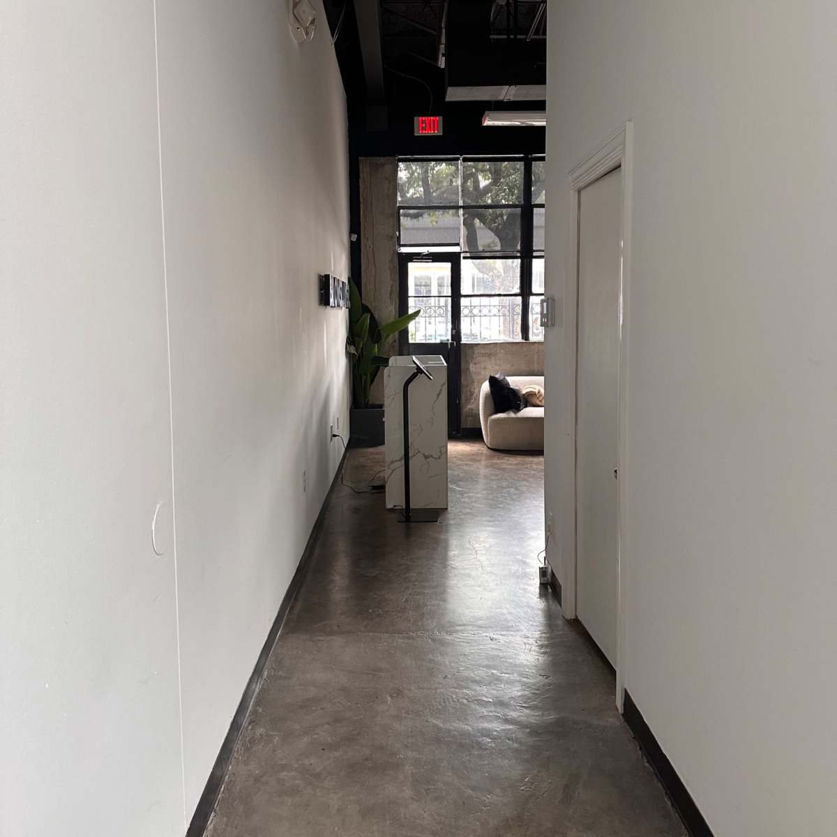 A narrow hallway with concrete flooring leads to a large window at the end, featuring a potted plant and a piece of furniture visible in the adjacent room.