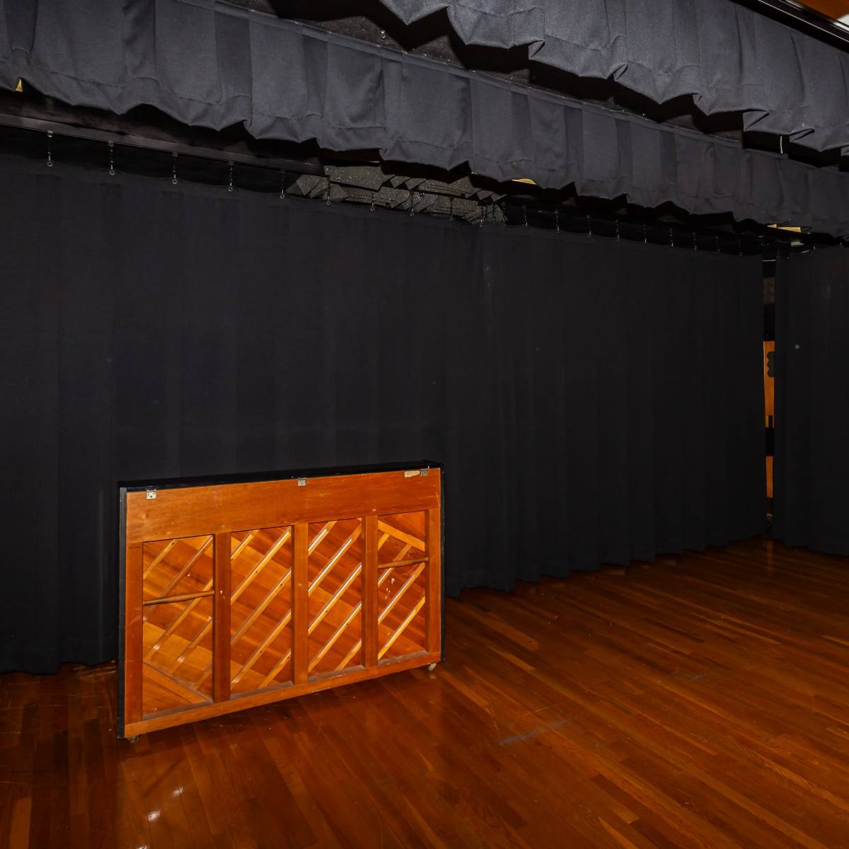 An empty stage with a wooden piano in front of a black curtain backdrop and polished wooden flooring.