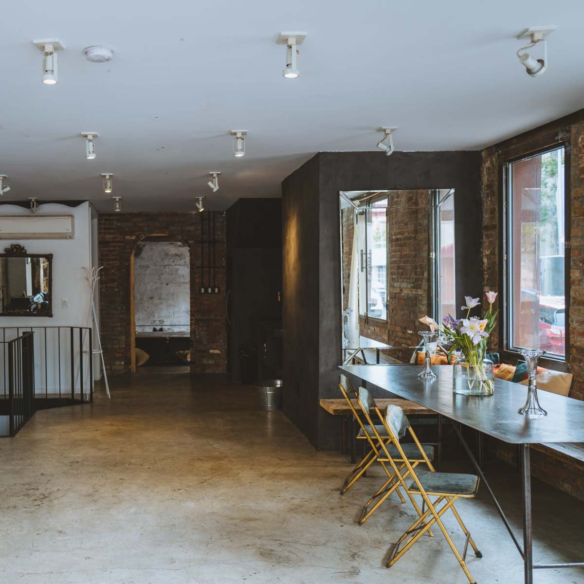 A spacious, modern interior with exposed brick walls, a black leather sofa, a metal dining table, and large windows allowing natural light to enter.