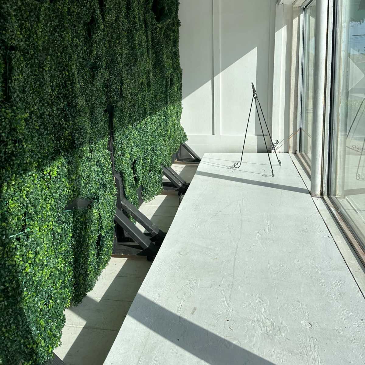 A sunlit indoor space featuring a green wall of artificial foliage alongside a long, empty table with a metal easel.