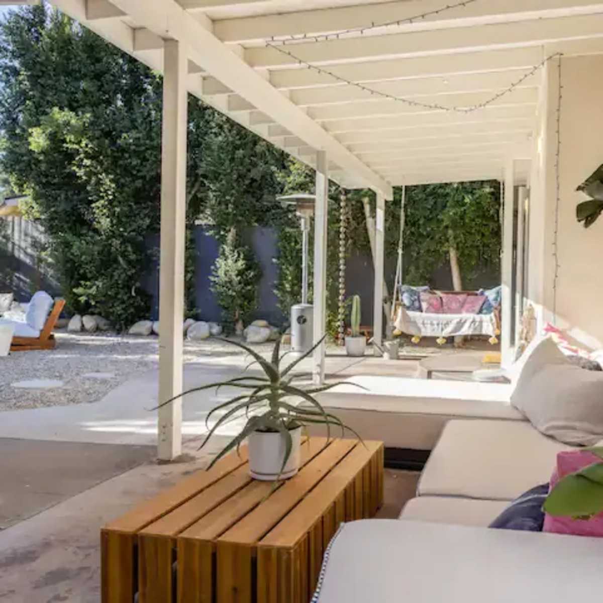 A covered patio area features seating, a wooden table, and lush greenery in the background.