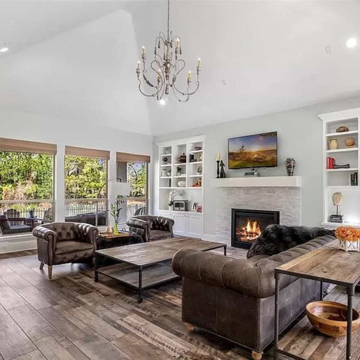 A modern living room with a fireplace, two leather sofas, a wooden coffee table, and large windows overlooking a green outdoor space.