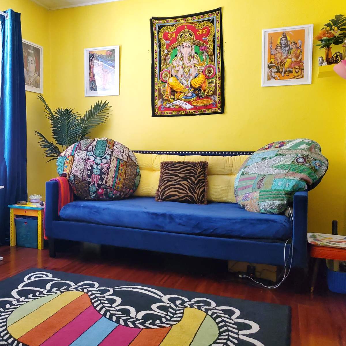 A bright yellow room features a blue sofa with patterned cushions, a colorful rug, and framed artwork on the walls.