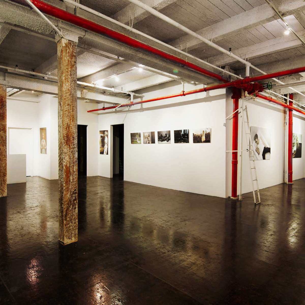 A spacious art gallery with polished dark floors, exposed ceiling beams, and white walls displaying several framed artworks.
