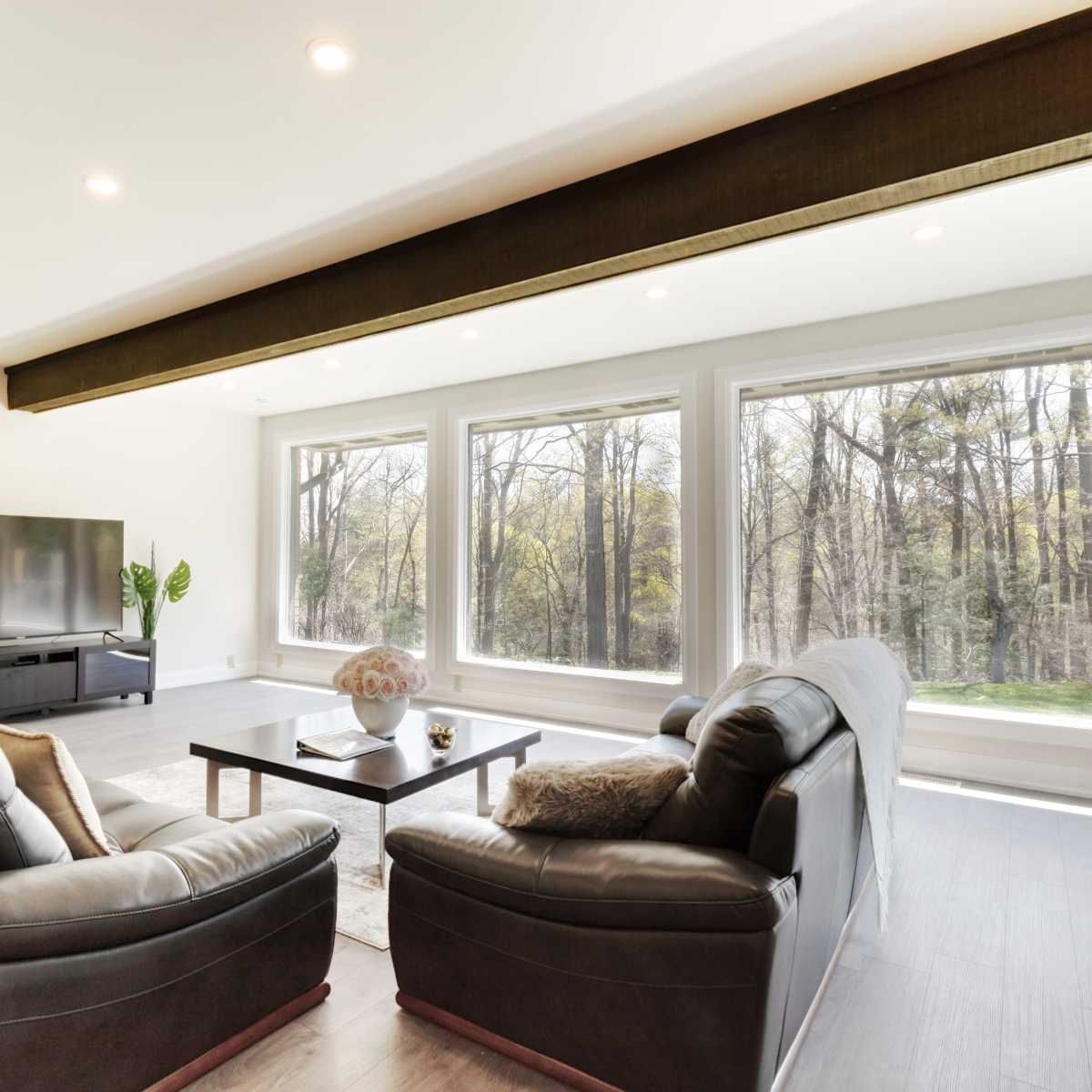 A modern living room features two leather sofas facing a coffee table, with a television and large windows showcasing a view of trees outside.