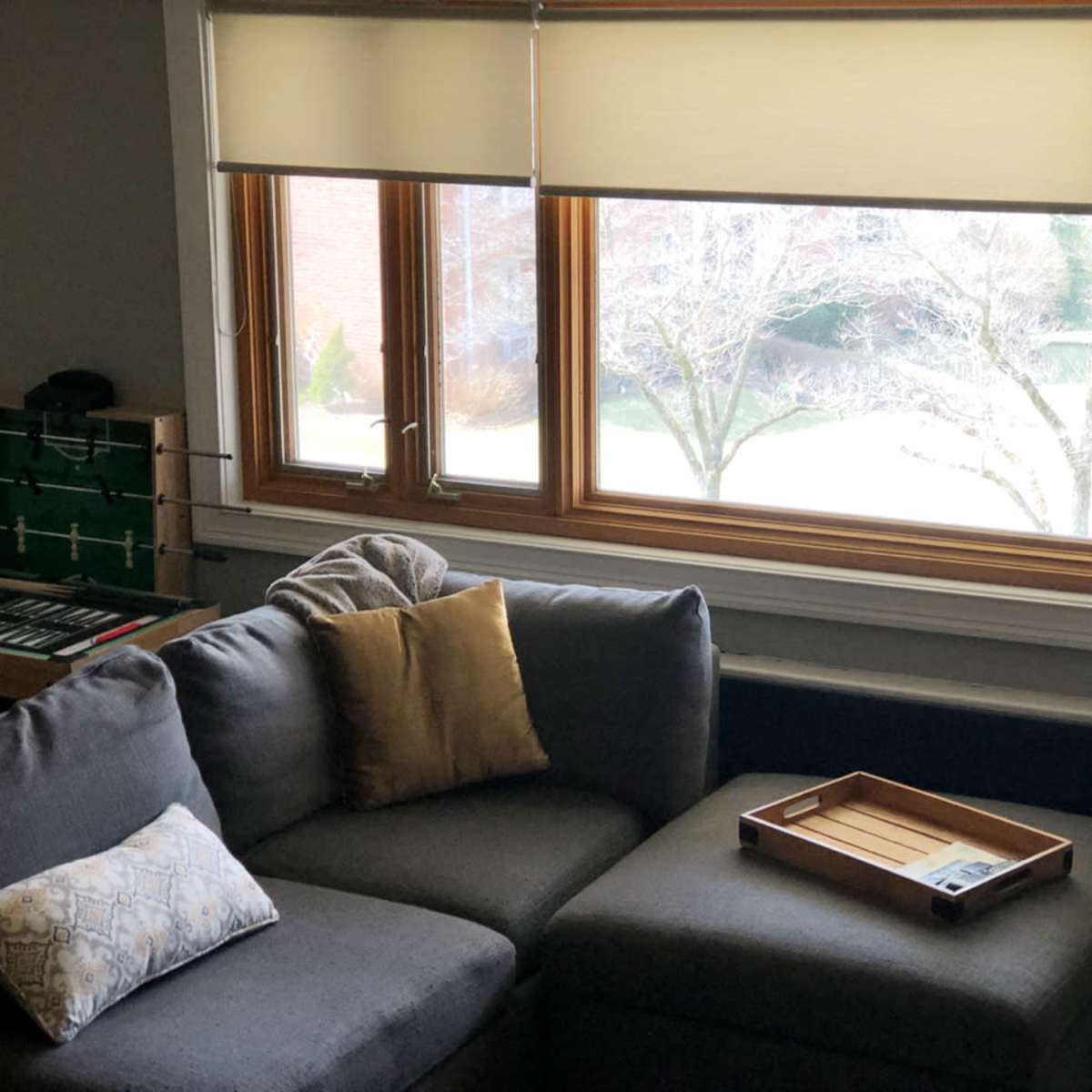 A cozy living room corner with a gray sectional sofa, decorative pillows, a wooden tray on the ottoman, and a window overlooking a yard with bare trees.