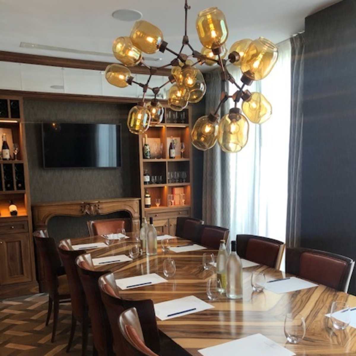 A long wooden dining table is set with glasses and notepads, illuminated by a modern chandelier, in a well-furnished room featuring a wall-mounted TV and shelves with bottles.
