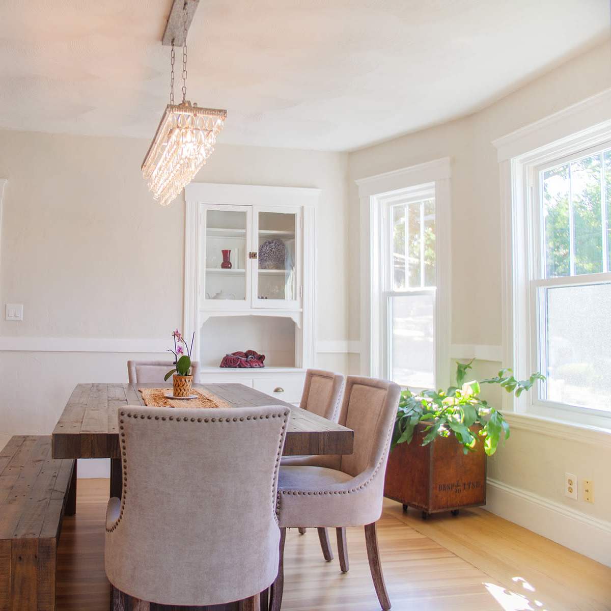 A bright dining room featuring a wooden table surrounded by plush chairs, with a modern chandelier above and a china cabinet against the wall.