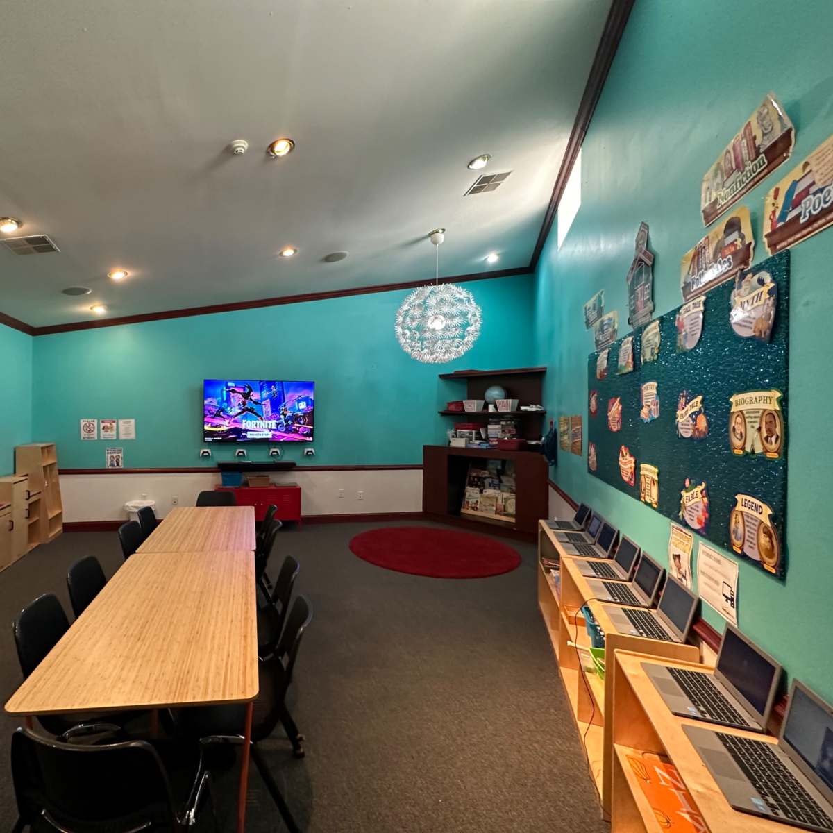 A brightly colored room featuring a long table, chairs, a wall-mounted TV, and several laptops arranged on a table along with some educational posters on the walls.