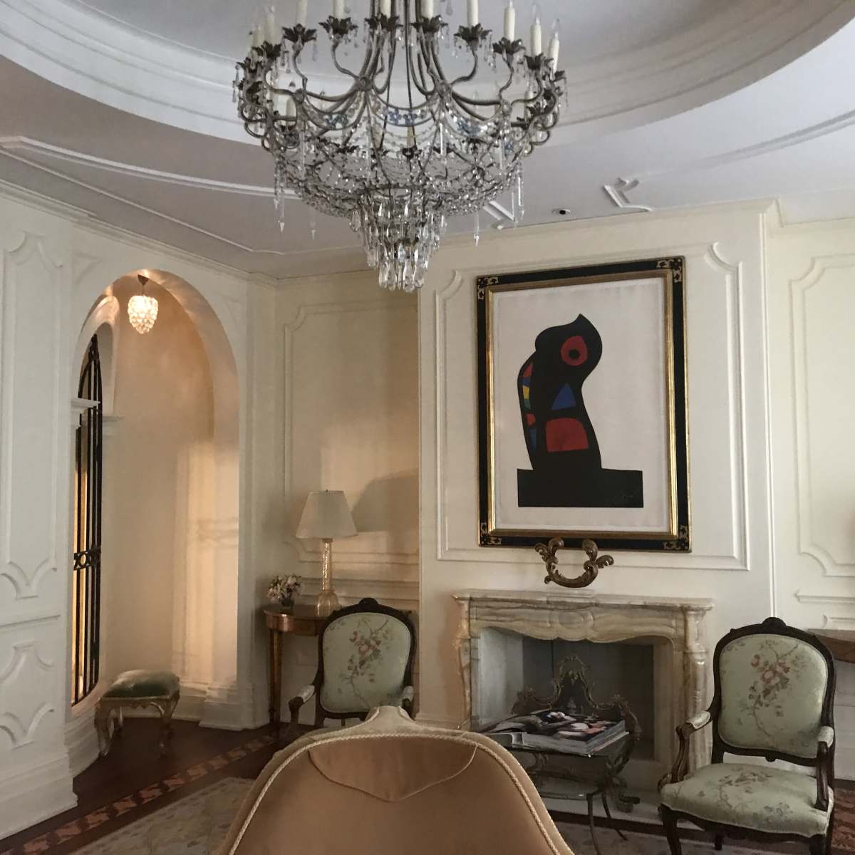 The room features a large chandelier, an ornate fireplace, two patterned chairs, and a colorful abstract painting on the wall.