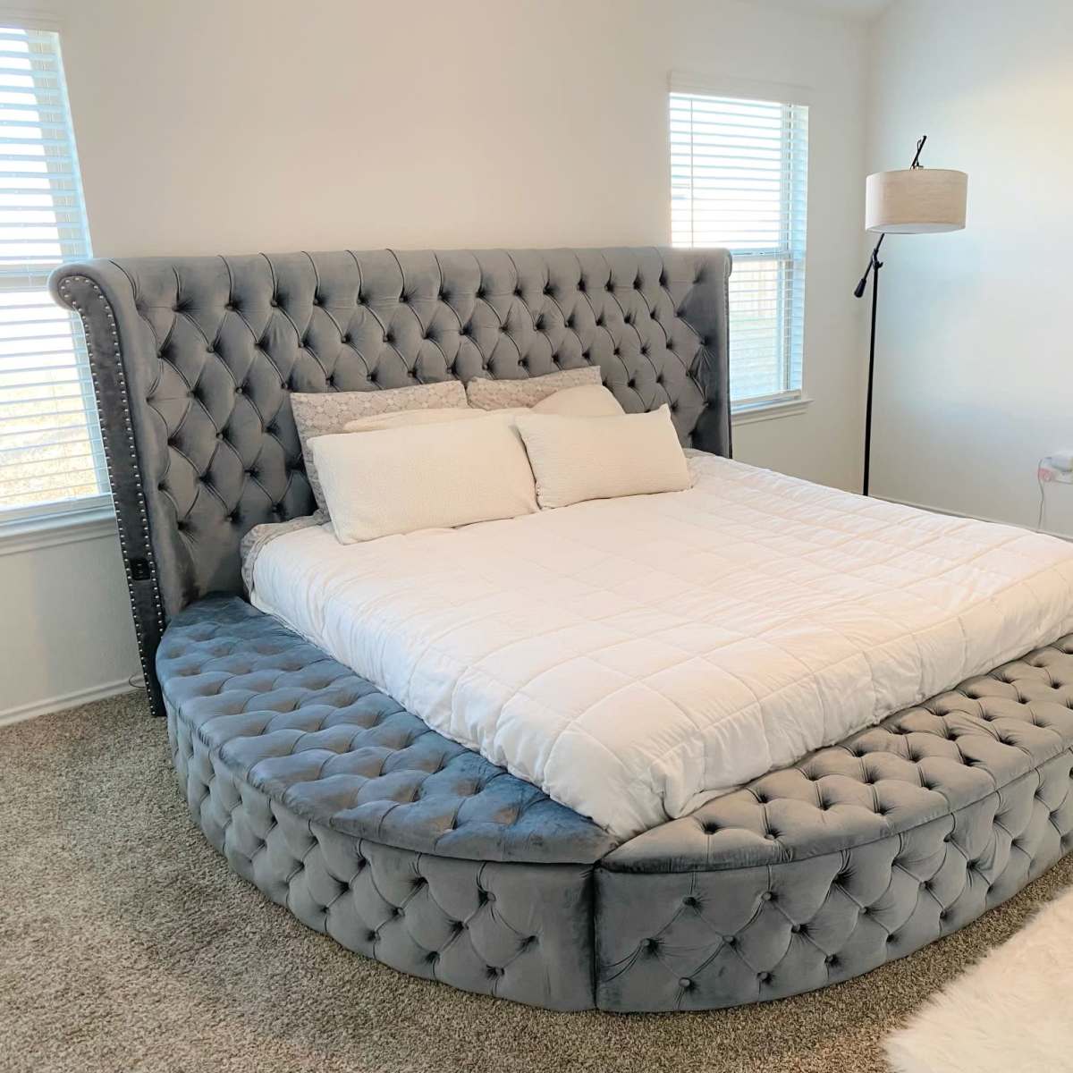 A round tufted bed with a gray fabric finish positioned in a well-lit bedroom with two windows and a floor lamp.