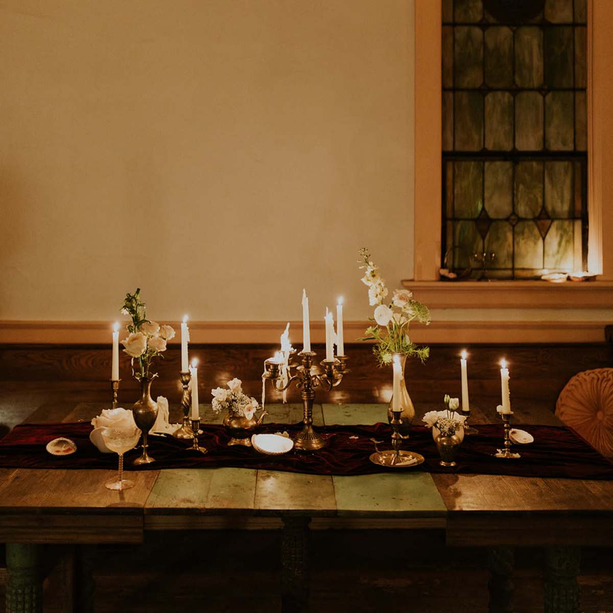 The scene features a dimly lit table set with candles, floral arrangements, and decorative dishes, positioned next to a stained glass window.