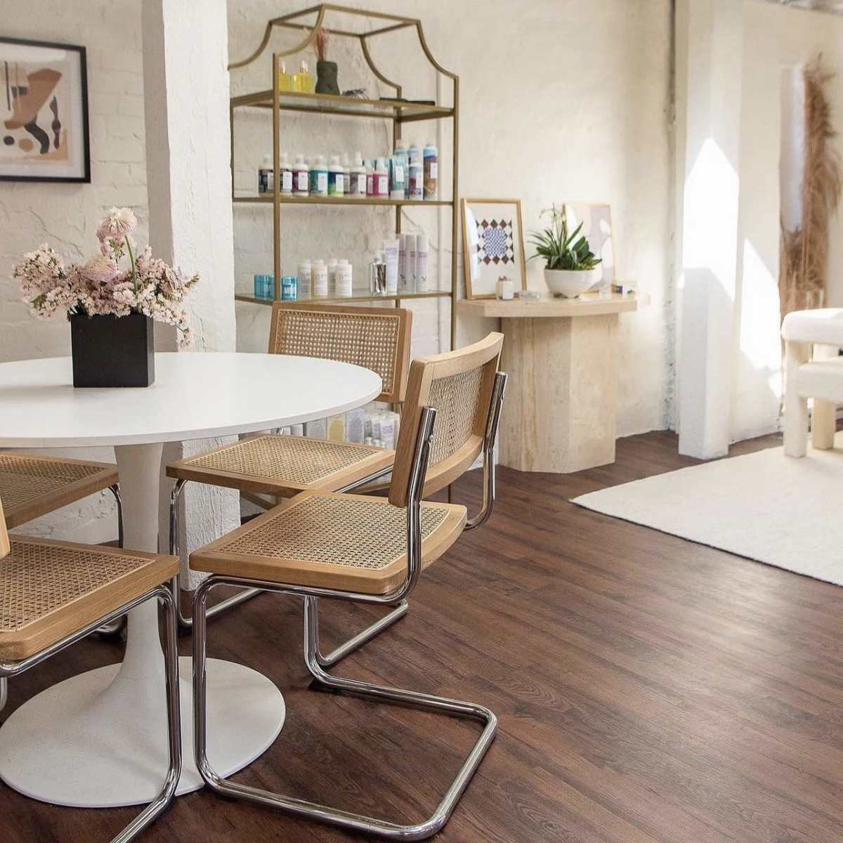 A bright, modern interior with a round white table surrounded by four cane-back chairs, alongside a decorative shelf displaying various bottles and plants.