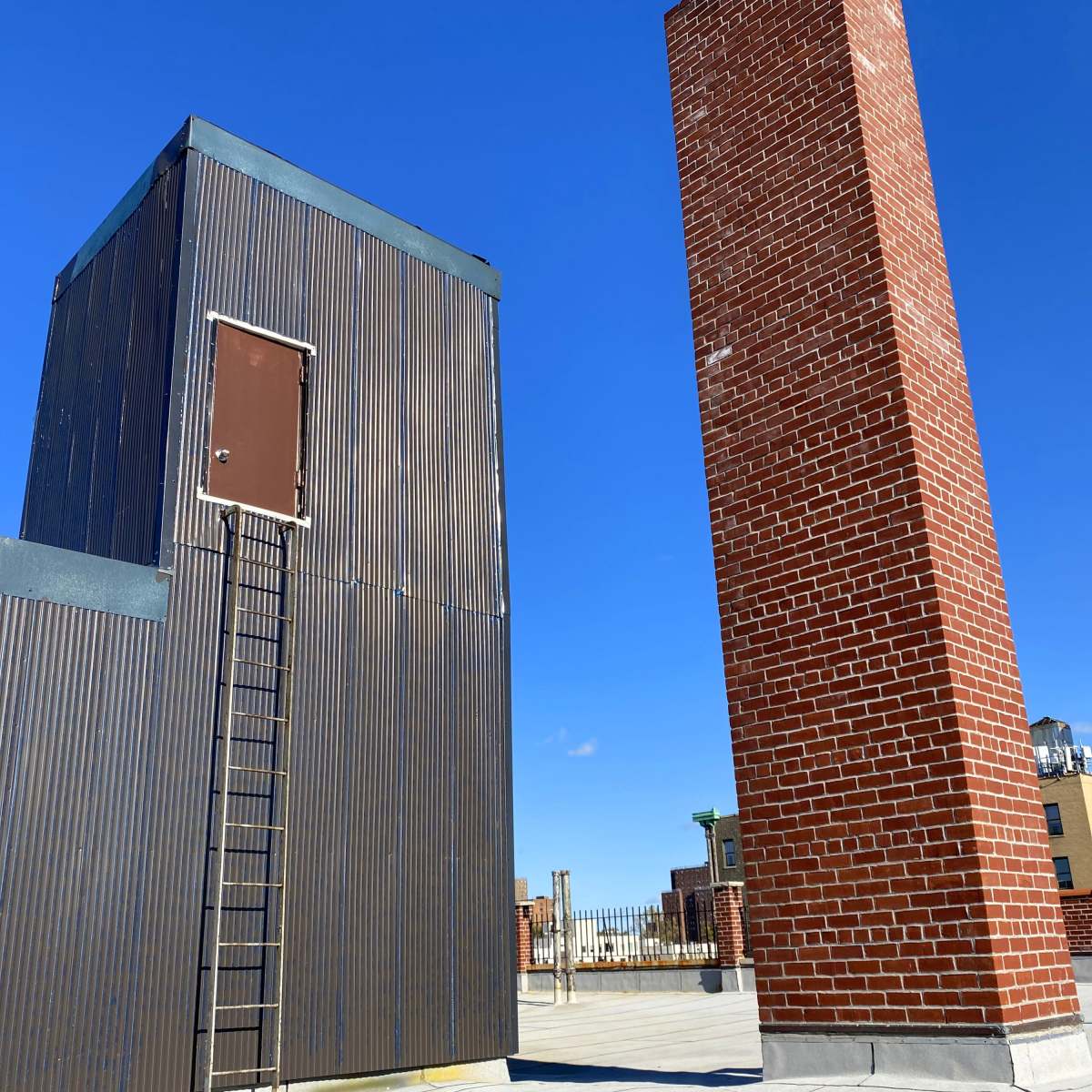A rooftop scene featuring a metal structure with a ladder leading to a door, alongside a tall brick chimney under a clear blue sky.