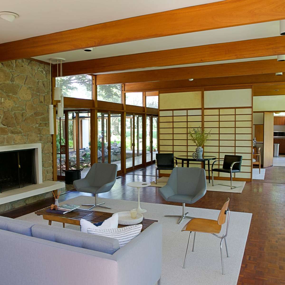 A modern living room with a stone fireplace, large windows, and minimalist furniture arranged around a central area rug.