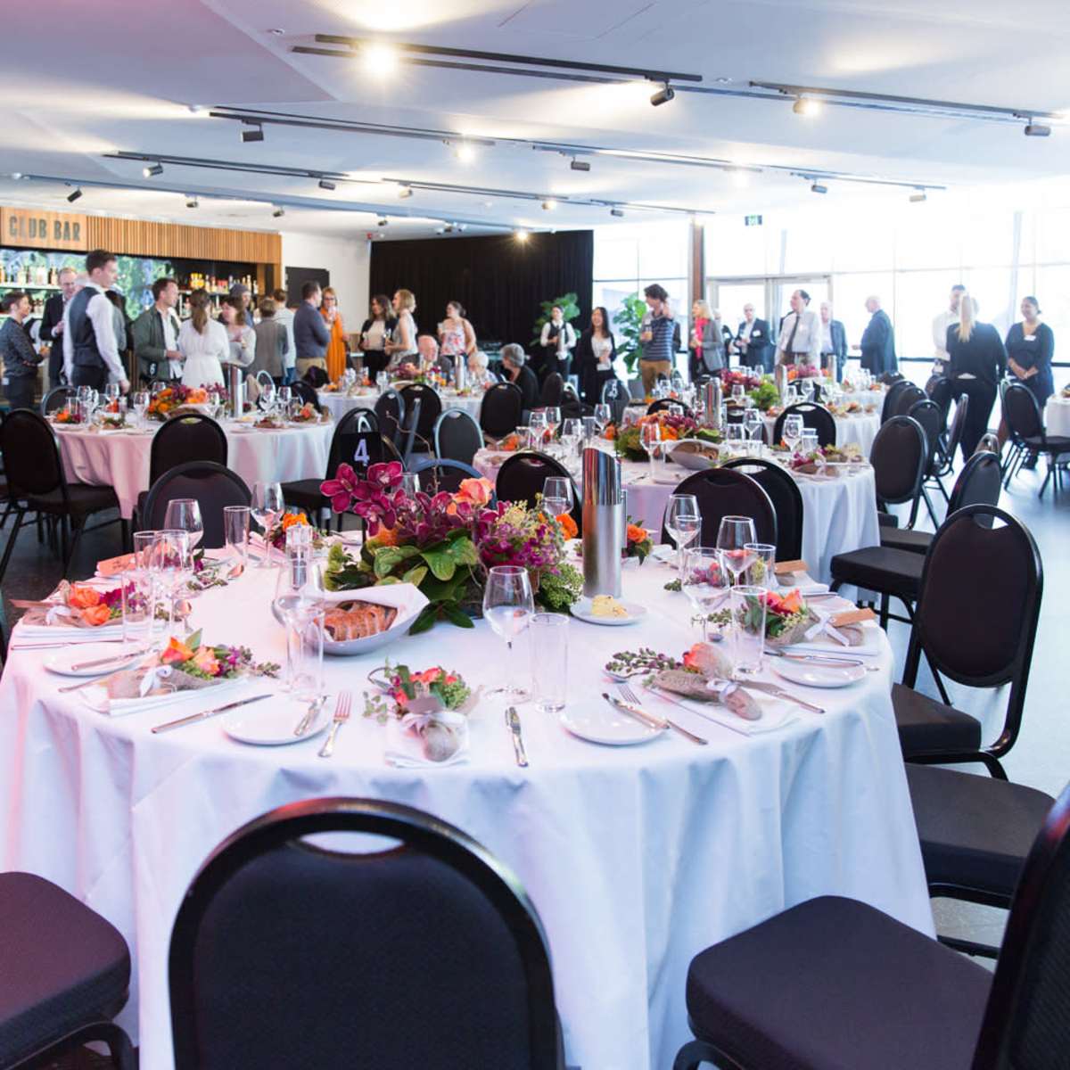 A banquet hall is set with elegantly arranged tables, featuring white tablecloths and floral centerpieces, while guests mingle in the background.