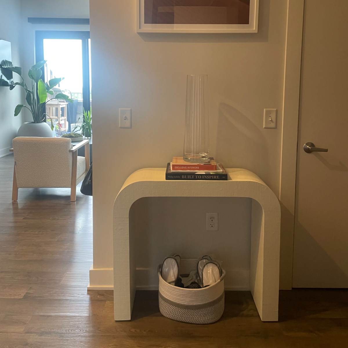 A curved white console table with a vase sits against a beige wall, with a framed artwork above it and a basket containing shoes underneath.