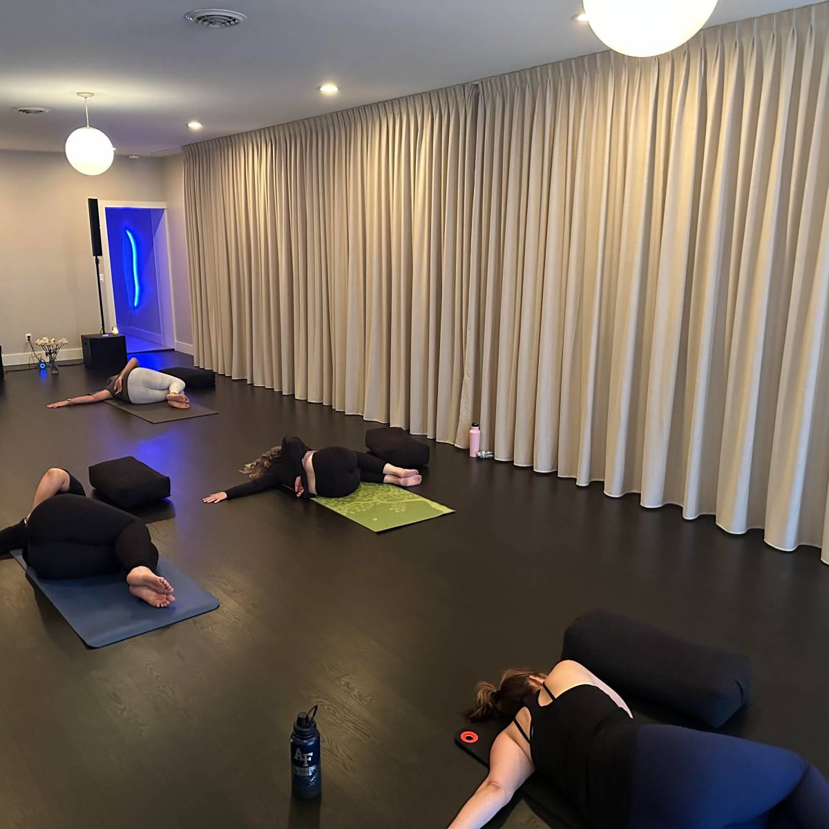 A group of individuals practice a yoga session on mats in a studio with soft lighting and beige curtains.